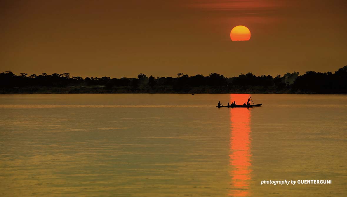 A boat is positioned in the reflection of the setting sun on a river.