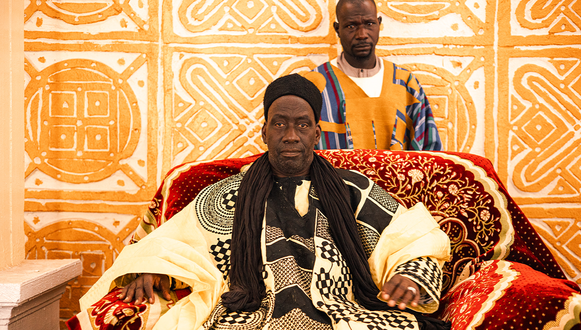 A king sits in his throne. Ornate patterns adorn his clothes, the red throne, and the wall behind.