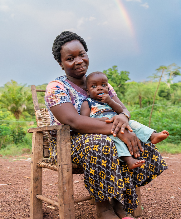 A woman holds her child in her lap in a wooden chair. Green rainforest and a rainbow are in the background.