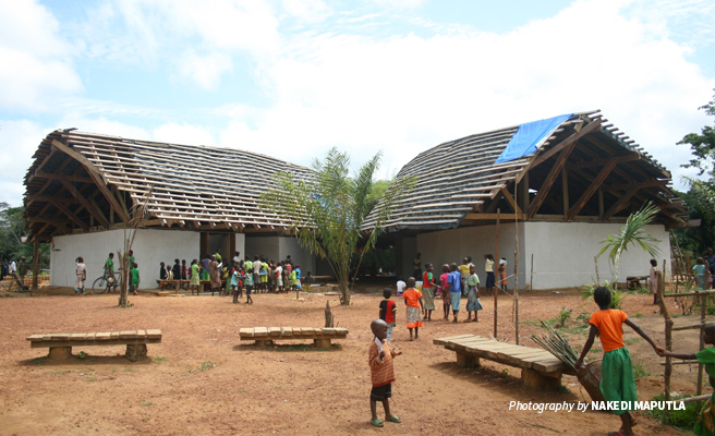 Photo of Ilima Conservation Primary School with untiled roof with children from the community in the foreground Photo of Ilima Conservation Primary School with untiled roof with children from the community in the foreground