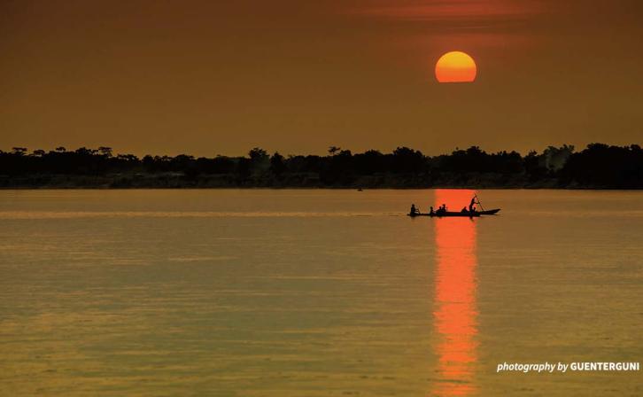 A boat is positioned in the reflection of the setting sun on a river.