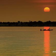 A boat is positioned in the reflection of the setting sun on a river.