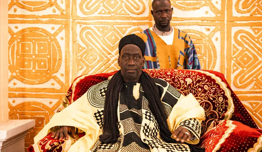 A king sits in his throne. Ornate patterns adorn his clothes, the red throne, and the wall behind.